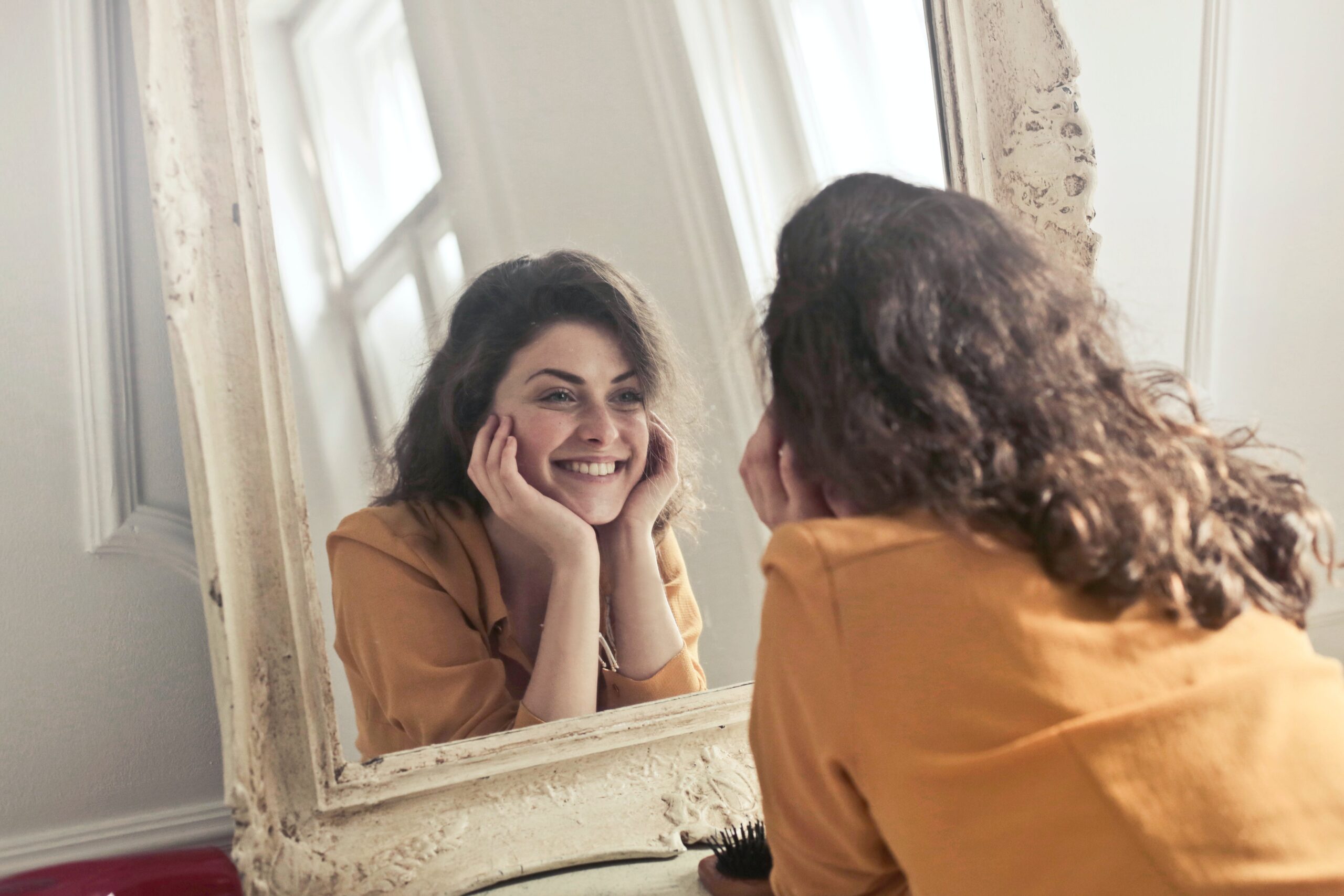 una giovane donna esprime gioia e fiducia mentre osserva il suo splendido sorriso riflettersi nello specchio, con le mani posate sul mento a sostegno del viso